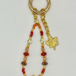 May include: A gold-colored keychain with a beaded charm. The charm features a mix of red, orange, and purple beads, along with gold-colored flower and star-shaped accents. A gold maple leaf charm hangs from a chain.