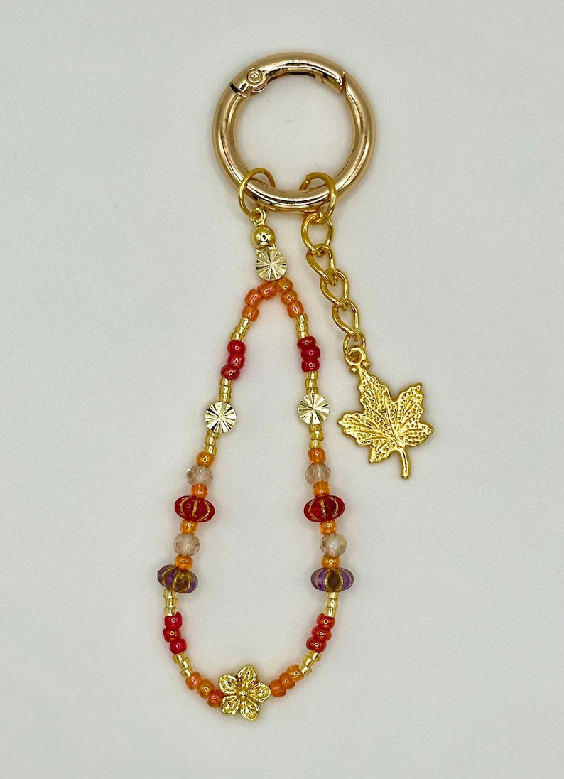 May include: A gold-colored keychain with a beaded charm. The charm features a mix of red, orange, and purple beads, along with gold-colored flower and star-shaped accents. A gold maple leaf charm hangs from a chain.