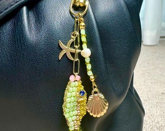 May include: A gold-toned keychain with a beaded fish, starfish, and seashell charm. The fish is green and gold, with a blue gem eye. The keychain is attached to a black leather bag.