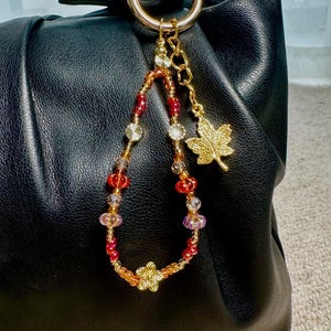 May include: A black leather handbag with a gold-tone ring and a beaded charm. The charm features red, orange, and clear beads, a gold flower, and a gold maple leaf charm. The bag has a gathered design.