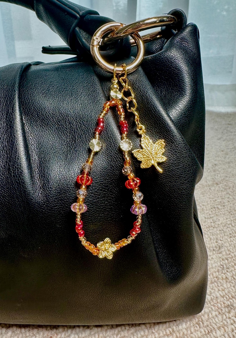 May include: A black leather handbag with a gold-tone ring and a beaded charm. The charm features red, orange, and clear beads, a gold flower, and a gold maple leaf charm. The bag has a gathered design.