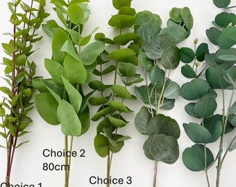 May include: Five artificial eucalyptus stems with varying leaf shapes and shades of green. Labels indicate Choice 1 at 97 cm, Choice 2 at 80 cm, Choice 3 at 84 cm, Choice 4 at 67 cm, and Choice 5 at 85 cm, showing distinct foliage textures and stem lengths.