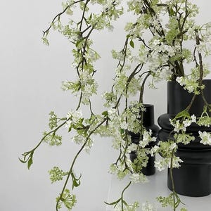 May include: Two matte black vases with a tiered, cylindrical design holding long, arching branches covered in small white blossoms and light green leaves. The branches drape gracefully over the dark, textured surfaces of the two containers.