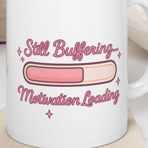 May include: White ceramic mug with the text "Still Buffering... Motivation Loading" in a pink script font. The design includes a pink progress bar. The mug sits on a beige coaster.