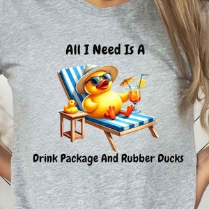 May include: Gray t-shirt with a graphic of a yellow rubber duck wearing sunglasses and a hat, relaxing on a beach chair with a drink. The text "All I Need Is A Drink Package And Rubber Ducks" is printed below the graphic.