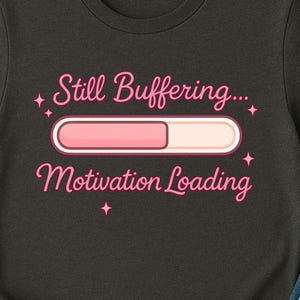 Puede incluir: Camiseta gris oscuro con el texto "Still Buffering... Motivation Loading" en rosa. El diseño incluye una barra de progreso rosa. La camiseta se combina con vaqueros azules y una taza blanca.