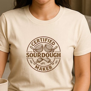 May include: A beige t-shirt with a round brown graphic that reads "CERTIFIED SOURDOUGH MAKER". The graphic features a loaf of bread and a rolling pin. The person is holding a beige mug.