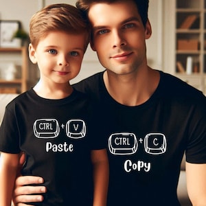 May include: A father and son in matching black t-shirts. The son's shirt features the text "CTRL + V Paste" in white, and the father's shirt says "CTRL + C Copy" in white. The image is a portrait of the two.