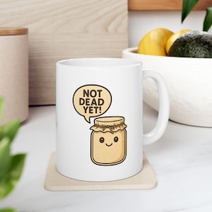 May include: White ceramic mug featuring a cartoon illustration of a jar with a smiling face and the text "NOT DEAD YET!" in a speech bubble. The jar is light brown with a lid and a cute design.