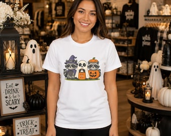 Op de afbeelding: Een wit t-shirt met korte mouwen en een afbeelding van twee wasberen en een geest. De ene wasbeer draagt een paarse heksenhoed en cape, de geest houdt een oranje snoepzakje vast en de andere wasbeer zit in een uitgeholde pompoen.