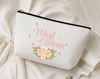 May include: A white cosmetic bag with a black zip and gold pull. The bag features the text "Maid of Honor" in a soft pink script, with floral and star graphics. The bag is made of a smooth fabric.