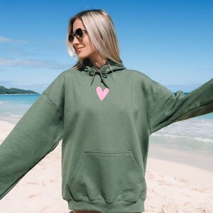 May include: A green hooded sweatshirt with a pink heart on the front. The sweatshirt is worn by a person standing on a beach.