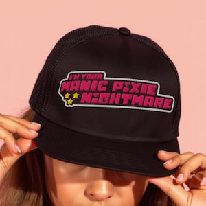 Puede incluir: Gorra de camionero negra con el texto "I'M YOUR MANIC PIXIE NIGHTMARE" en rosa y blanco. La gorra tiene una parte trasera de malla y una visera curva. El texto está en una fuente audaz y estilizada con estrellas.