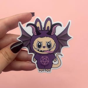 May include: A purple sticker featuring a cartoon demon character with bat wings, horns, and a pentagram on its chest. The character has a mischievous expression with its tongue sticking out. The sticker is held against a pink background.