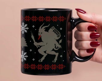 Puede incluir: Taza de cerámica negra con un diseño de Krampus. La taza tiene un asa negra y un patrón repetitivo de copos de nieve rojos y diseños florales. La ilustración de Krampus es gris y sostiene un haz de ramas de abedul.