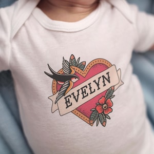 May include: White baby bodysuit featuring a heart-shaped tattoo design. The design includes a swallow, a banner with the name "EVELYN", and a red flower. The bodysuit is short-sleeved and made of soft material.