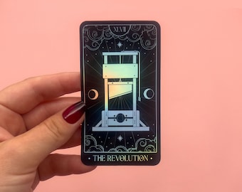 May include: A rectangular sticker featuring a guillotine illustration on a black background. The design includes celestial elements like stars and clouds, with the text "THE REVOLUTION" at the bottom. The sticker has a holographic effect.