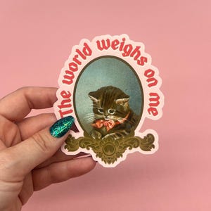 May include: A sticker with a vintage-style illustration of a kitten wearing a bow. The kitten is inside an oval frame with the words "The world weighs on me" in red text. The sticker has a pink background.