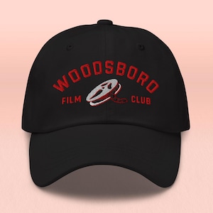 May include: Black baseball cap with red embroidered text reading "WOODSBORO FILM CLUB". The cap features a film reel graphic. A classic, adjustable hat for film enthusiasts.