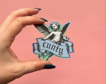 May include: A sticker featuring a cherubic angel with wings, holding a banner with the word "c*nty" in a gothic font. The sticker has a holographic, iridescent border and is held against a pink background.