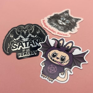 May include: Three stickers on a pink background. One features a black illustration of Baphomet with the text "SATAN IS MY HOMEBOY." Another shows a cat with the text "Perhaps today, Satan." The third is a cartoon demon.