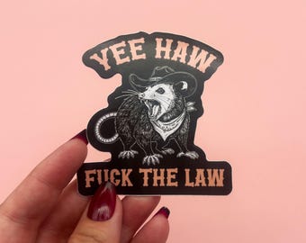 May include: A black sticker featuring a cartoon opossum wearing a cowboy hat and bandana. The text "YEE HAW" is above the opossum, and "FUCK THE LAW" is below. The sticker is held against a pink background.