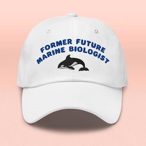 Può includere: Cappellino da baseball bianco con la scritta blu "FORMER FUTURE MARINE BIOLOGIST" arcuata sopra un'illustrazione di un'orca in bianco e nero. Il cappellino ha una visiera curva e una chiusura regolabile.