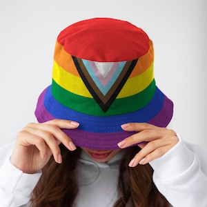 Może przedstawiać: Kapelusz typu bucket z tęczowym wzorem, na którym znajduje się czarny trójkąt z brązowymi, białymi i różowymi paskami. Kapelusz wykonany jest z miękkiej, wygodnej tkaniny i idealnie nadaje się do codziennego noszenia.