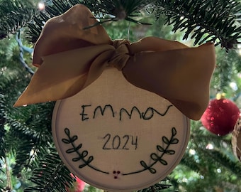 May include: A white, round, embroidered Christmas ornament with a brown ribbon. The ornament says "Emma 2024" in black embroidery.