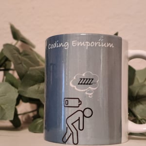 May include: A white ceramic mug with a blue and gray ombre design. The mug has the text "coding Emporium" printed on the front. A cartoon stick figure is depicted with a thought bubble above its head that says "ZZZZ..." and a low battery symbol next to it.