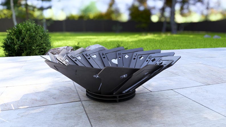 May include: A black metal fire pit with a unique design featuring playing card symbols. The fire pit is set on a tiled patio with a grassy background, perfect for outdoor gatherings.