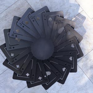May include: A circular arrangement of black playing cards fanned out from a central dark gray disc. The cards display suits of hearts, diamonds, clubs, and spades. The background is a light gray tiled surface.