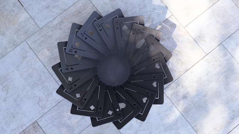 May include: A circular arrangement of black playing cards fanned out from a central dark gray disc. The cards display suits of hearts, diamonds, clubs, and spades. The background is a light gray tiled surface.