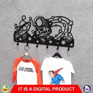 Op de afbeelding: Zwarte metalen wandkapstok met ruimtethema: astronaut, raket, sterren en planeten. Daaronder hangen een oranje en wit shirt met de tekst "ASK MORE QUESTIONS" en een wit shirt met een Superman-afbeelding. De tekst "IT IS A DIGITAL PRODUCT" staat onderaan.