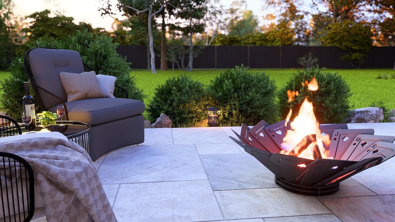 May include: A backyard patio scene featuring a modern fire pit with playing card cutouts, flames, and a gray lounge chair with pillows. A small table with a bottle of wine and a blanket complete the outdoor setting.