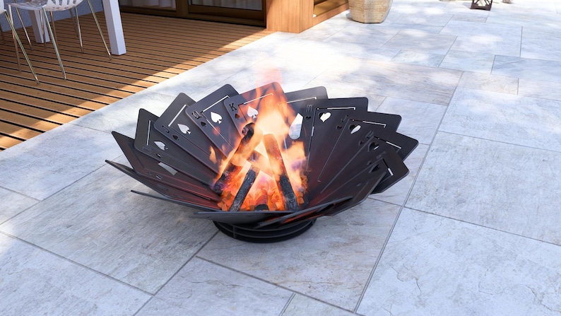 May include: A dark metal fire pit with a unique design featuring playing card symbols. The fire pit is filled with burning wood, producing bright orange flames. The fire pit is sitting on a light gray tiled surface.