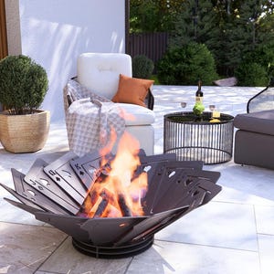 May include: A modern outdoor patio scene featuring a decorative fire pit with a playing card design, flames, and comfortable seating. The scene includes a round side table, a potted plant, and a bottle of wine.