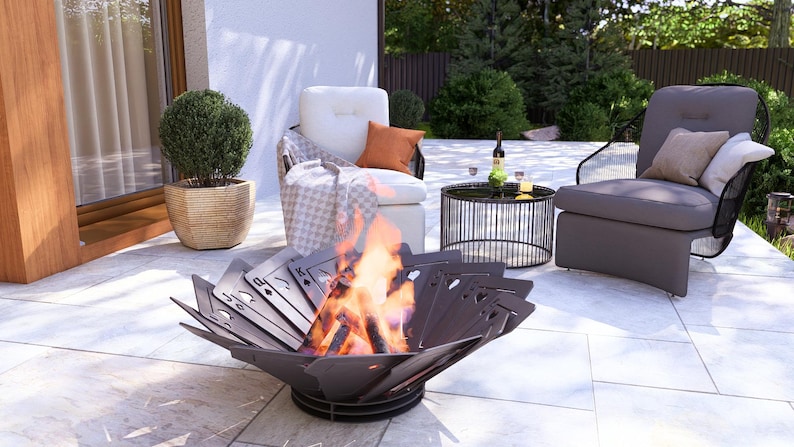 May include: A modern outdoor patio scene featuring a decorative fire pit with a playing card design, flames, and comfortable seating. The scene includes a round side table, a potted plant, and a bottle of wine.