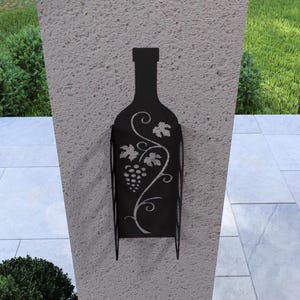 Puede incluir: Porta botellas de vino de metal negro con un dise&ntilde;o de vid y uvas. El soporte est&aacute; montado en una pared gris claro.