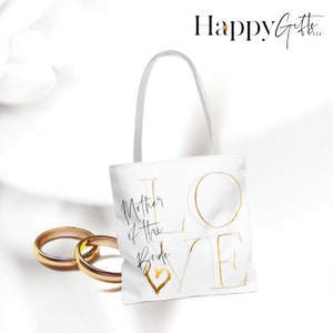 Puede incluir: Bolsa de tela blanca con letras doradas que dicen "LOVE" y "Mother of the Bride" con un coraz&oacute;n dorado.