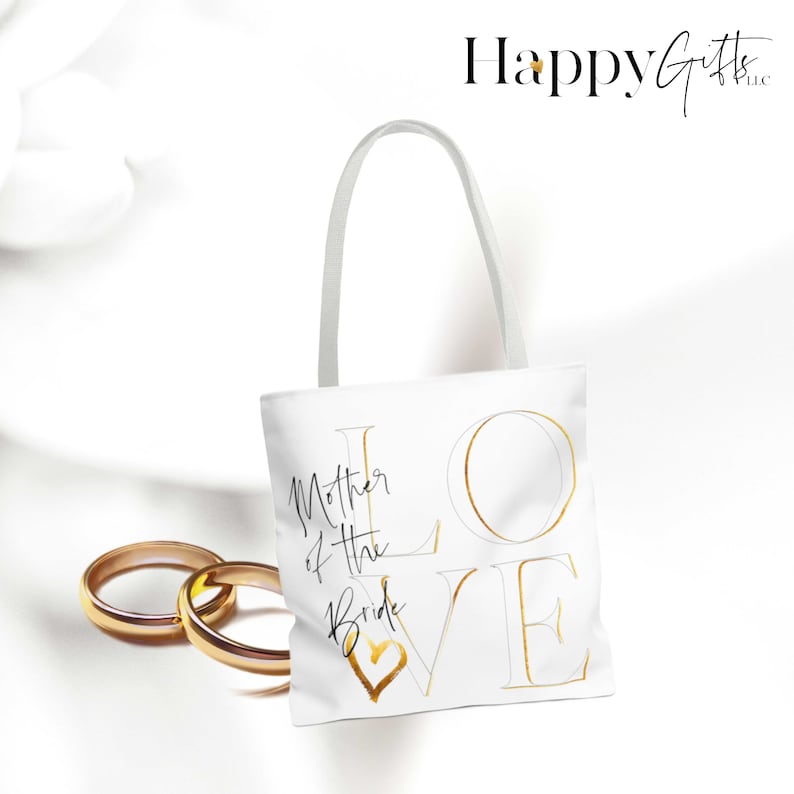 Puede incluir: Bolsa de tela blanca con letras doradas que dicen "LOVE" y "Mother of the Bride" con un coraz&oacute;n dorado.