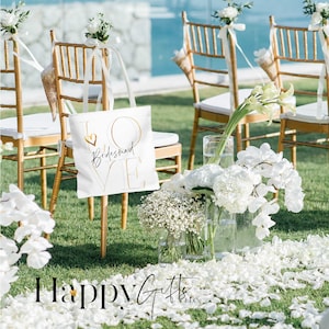 Puede incluir: Bolsa de tela blanca con letras doradas que dicen "LOVE Bridesmaid" con un peque&ntilde;o coraz&oacute;n en la "O". La bolsa est&aacute; sentada en una silla dorada en un entorno al aire libre con flores blancas y p&eacute;talos en el suelo.