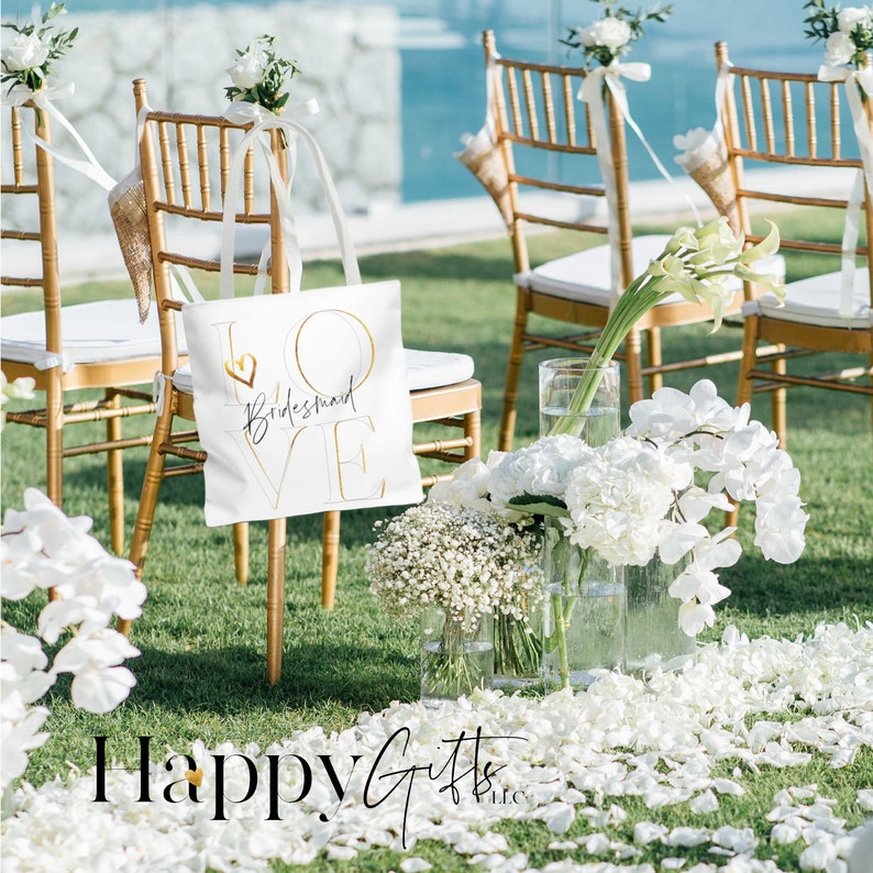 Puede incluir: Bolsa de tela blanca con letras doradas que dicen "LOVE Bridesmaid" con un peque&ntilde;o coraz&oacute;n en la "O". La bolsa est&aacute; sentada en una silla dorada en un entorno al aire libre con flores blancas y p&eacute;talos en el suelo.