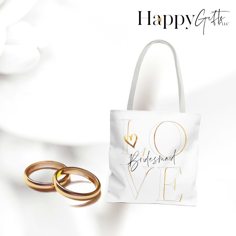 Puede incluir: Bolsa de tela blanca con letras doradas que dicen "LOVE" con un peque&ntilde;o coraz&oacute;n en la "O". La palabra "bridesmaid" est&aacute; escrita en cursiva negra debajo del "LOVE".
