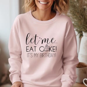 以下が含まれることがあります： 「let me... EAT CAKE! IT'S MY BIRTHDAY!」の黒い筆記体テキストが特徴の、明るいピンク色のスウェットシャツ。小さなケーキのグラフィックが含まれています。スウェットシャツはカジュアルなスタイルです。