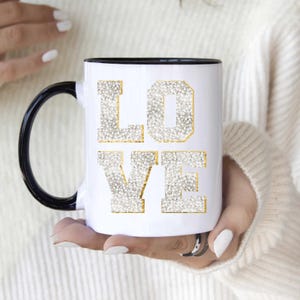May include: White ceramic mug with a black rim and handle. The mug features the word "LOVE" in gold lettering with a white, sparkly background.