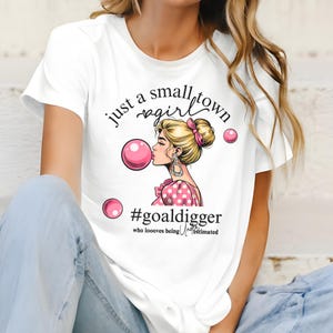 Può includere: Maglietta bianca con una grafica rosa e nera di una donna che soffia una bolla con il testo "just a small town girl #goaldigger who loooves being underestimated"