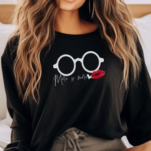 May include: Black sweatshirt with a white graphic of a pair of glasses and a red lip print. The text "More is more" is printed below the glasses.