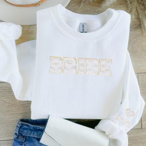 May include: White sweatshirt with the word "BRIDE" in gold glitter lettering. The sweatshirt has a small heart with the word "Love" on the sleeve.