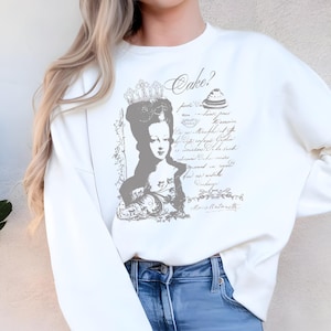 Op de afbeelding: Crèmekleurige sweatshirt met een grijze afbeelding van Marie Antoinette, een kroon en de tekst "Cake?". Het ontwerp bevat script en een taartillustratie. De sweatshirt heeft lange mouwen en een ronde hals.
